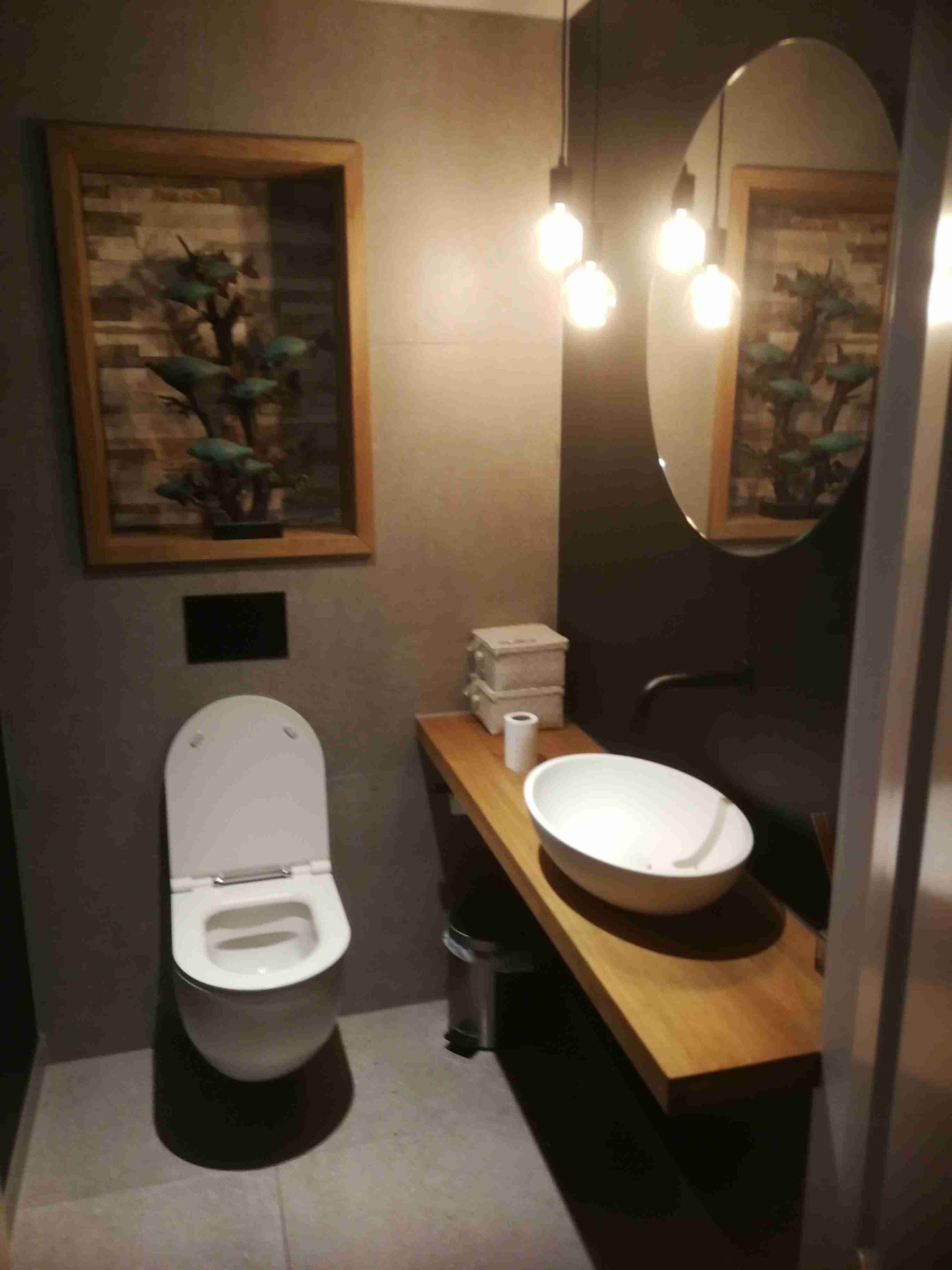 Donkere toiletruimte met hangend toilet, houten plank met waskom en ronde spiegel